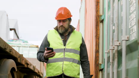 Worker-Wearing-Vest-And-Safety-Helmet-Reading-On-A-Smartphone-And-Walking-In-A-Logistics-Park
