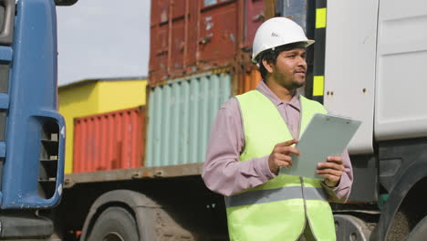 Worker-Wearing-Vest-And-Safety-Helmet-Looking-And-Smiling-At-Camera-In-A-Logistics-Park-While-Holding-Documents