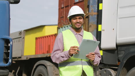 Worker-Wearing-Vest-And-Safety-Helmet-Looking-And-Smiling-At-Camera-In-A-Logistics-Park-While-Holding-Documents-1