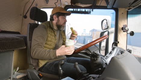 Side-View-Of-Worker-Sitting-In-A-Truck-In-A-Logistics-Park-While-Eating-A-Sandwitch-And-Holding-A-Drink