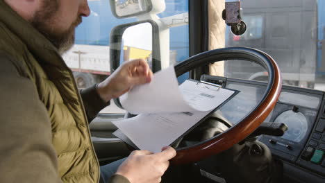 Rear-View-Of-Worker-Sitting-In-A-Truck-In-A-Logistics-Park-While-Reading-Documents