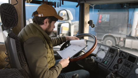 Side-View-Of-Worker-Sitting-In-A-Truck-In-A-Logistics-Park-While-Reading-Documents