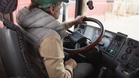 Rear-View-Of-Older-Worker-Wearing-Cap-And-Vest-Driving-A-Truck-In-A-Logistics-Park