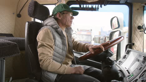 Side-View-Of-Older-Worker-Wearing-Cap-And-Vest-Driving-A-Truck-In-A-Logistics-Park