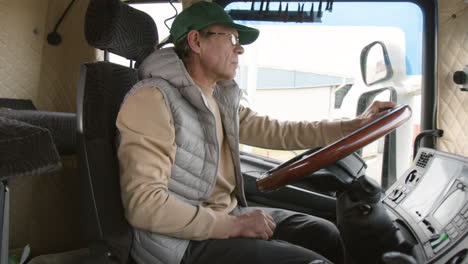 Side-View-Of-Older-Worker-Wearing-Cap-And-Vest-Driving-A-Truck-In-A-Logistics-Park-1