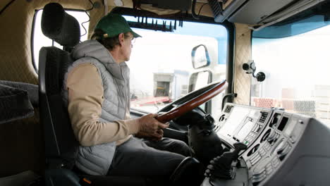 Side-View-Of-Older-Worker-Wearing-Cap-And-Vest-Driving-A-Truck-In-A-Logistics-Park-2