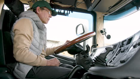 Side-View-Of-Older-Worker-Wearing-Cap-And-Vest-Driving-A-Truck-In-A-Logistics-Park-4