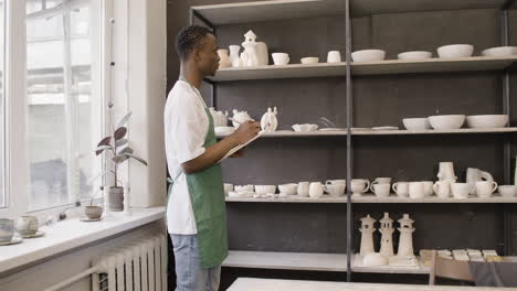 Young-American-Male-Clerk-Writing-On-A-Clipboard-And-Making-An-Inventory-Of-Ceramics-In-The-Pottery-Shop
