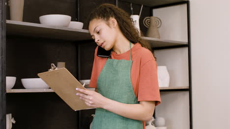 Young-Female-Clerk-Having-A-Call-And-Writing-On-A-Clipboard-In-The-Pottery-Shop