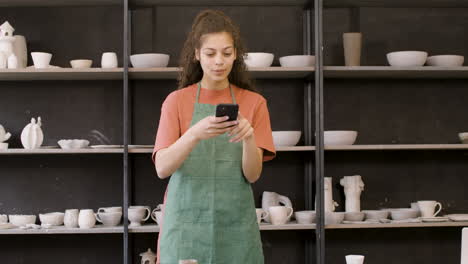 Woman-In-Apron-Using-Phone-And-Taking-Photos-Of-Handmade-Ceramic-Pieces-On-Table-In-The-Pottery-Shop