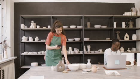 Woman-Taking-Photos-Of-Handmade-Ceramic-Pieces-And-Showing-Pictures-To-Her-Male-Colleague-Who-Working-On-Laptop-Computer-In-The-Pottery-Shop