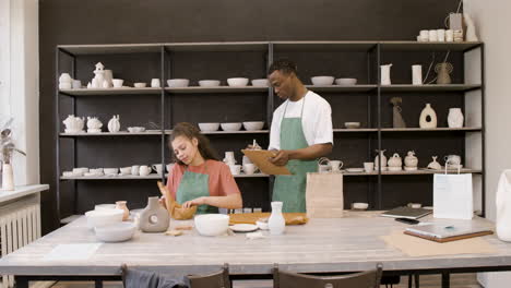 Young-Woman-Wrapping-Ceramic-Bowl-With-Paper-While-Her-Male-Colleague-Writing-On-Clipboard-And-Then-Putting-The-Bowl-In-A-Shopping-Bag-At-The-Pottery-Store