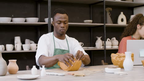 Young-Man-Packaging-Handicraft-Ceramics-With-Paper-In-The-Pottery-Shop-While-His-Female-Colleague-Working-On-Laptop-Computer-1