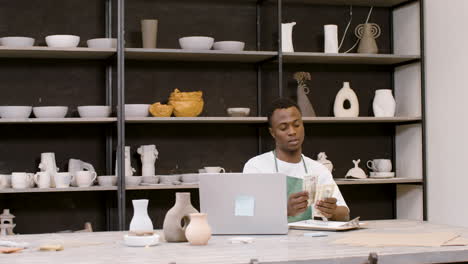 American-Clerk-Sitting-At-Desktop-In-Front-Of-Laptop-Counting-Banknotes-In-A-Pottery-Shop