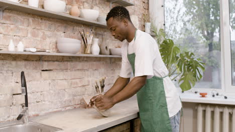 American-Clerk-In-Agreen-Apron-Kneading-Clay-On-Top-Of-A-Table-In-A-Pottery-Workshop