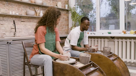 Employees-Wearing-Green-Apron-Modeling-Ceramic-Pieces-On-Potter-Wheel-In-A-Workshop-While-Talking-To-Each-Other-1