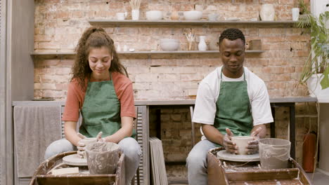 Front-View-Of-Employees-Wearing-Green-Apron-Modeling-Ceramic-Pieces-On-Potter-Wheel-In-A-Workshop-While-Talking-To-Each-Other-2