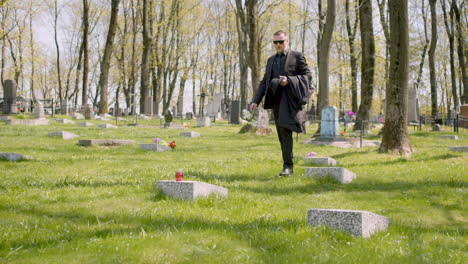 Sad-Man-In-Black-Suit-Walking-In-A-Graveyard,-Taking-Sunglassess-Off-And-Placing-A-White-Flower-On-A-Tombstone