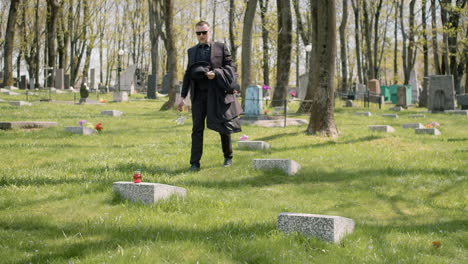 Sad-Man-In-Black-Suit-Walking-In-A-Graveyard,-Taking-Sunglassess-Off-And-Placing-A-White-Flower-On-A-Tombstone-1