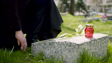 Close-Up-Of-An-Unrecognizable-Man-In-Black-Suit-Kneeling-And-Placing-A-White-Flower-On-A-Tombstone-In-A-Graveyard-1
