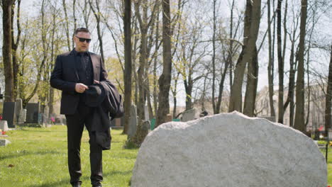 Sad-Man-In-Black-Suit-Walking-In-A-Graveyard,-Taking-Sunglasses-Off-And-Looking-At-A-Tombstone-Sorrowfully