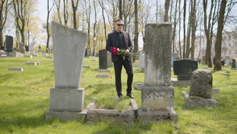Sad-Man-In-Black-Suit-Holding-Red-Roses-And-Walking-In-A-Graveyard,-Then-Taking-Sunglasses-Off-And-Looking-At-Tombstones-Sorrowfully
