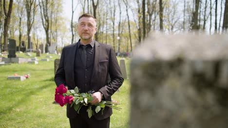 Sad-Man-In-Black-Suit-Standing-In-A-Graveyard-Holding-Red-Roses-While-Looking-At-The-Sky-And-At-Tombstones-Sorrowfully