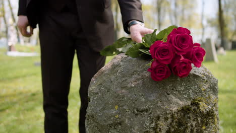 Close-Up-Of-An-Unrecognizable-Man-In-Black-Suit-Walking-And-Placing-Red-Roses-On-A-Tombstone-In-A-Graveyard
