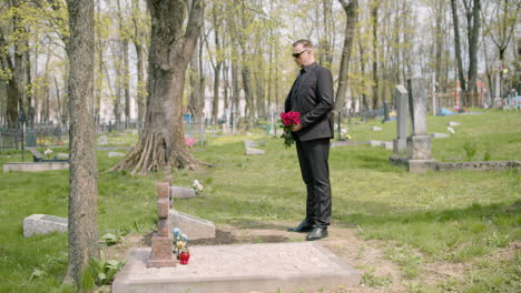 Man-In-Black-Suit-Holding-Red-Roses-Standing-In-Front-Of-A-Grave-In-A-Graveyard