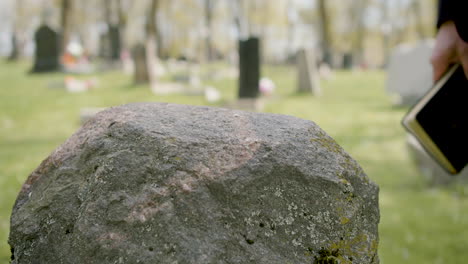 Close-Up-View-Of-Man-Hand-Touching-A-Tombstone-While-Holding-A-Bible-In-A-Graveyard