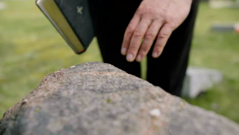 Close-Up-View-Of-Man-Hand-Touching-A-Tombstone-While-Holding-A-Bible-In-A-Graveyard-1