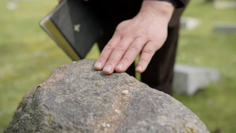 Close-Up-View-Of-Man-Hand-Touching-A-Tombstone-While-Holding-A-Bible-In-A-Graveyard-2