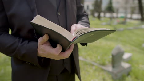 Close-Up-View-Of-Man-Hands-Holding-A-Bible-And-Reading-In-Front-Of-A-Tombstone-In-A-Graveyard