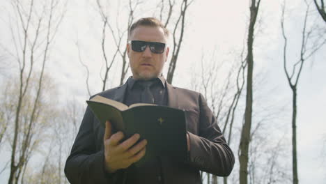 Unteransicht-Eines-Mannes-In-Schwarzem-Anzug-Und-Sonnenbrille,-Der-Auf-Einem-Friedhof-Eine-Bibel-Liest