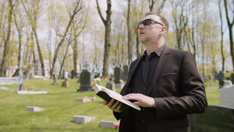Man-In-Black-Suit-And-Sunglasses-Reading-A-Bible-In-A-Graveyard