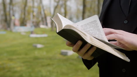 Close-Up-View-Of-Man-Hands-Holding-A-Bible-And-Reading-In-Front-Of-A-Tombstone-In-A-Graveyard-1