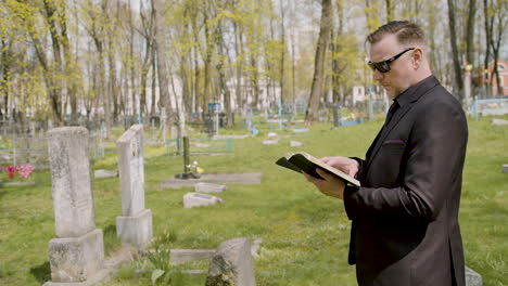 Man-In-Black-Suit-And-Sunglasses-Reading-A-Bible-In-Front-Of-A-Tombstone-In-A-Graveyard