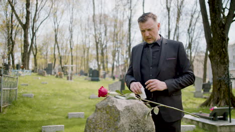 Man-In-Black-Suit-In-Front-A-Tombstone-Wiht-Red-Rose-While-He-Drying-His-Tears-And-Then-He-Leaves-The-Graveyard