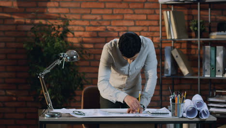 Young-Architect-Drawing-Some-Plan-On-The-Big-Sheet-Of-Paper-On-Desk-In-His-Office,-Then-He-Rises-Up-His-Head-And-Smiles-To-Camera