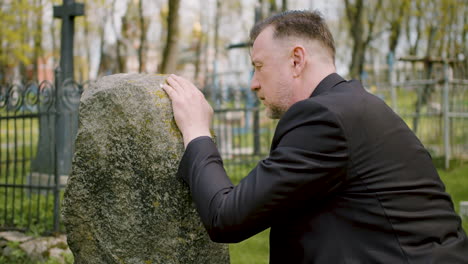 Close-Up-View-Of-Man-In-Black-Suit-Kneeling-In-Front-Of-A-Tombstone-And-Touching-It-1