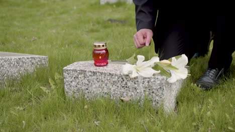 Unrecognizable-Man-In-Black-Suit-Kneeling-And-Putting-Flowers-And-A-Candle-On-Tombstone-In-A-Graveyard