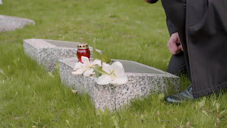 Unrecognizable-Man-In-Black-Suit-Kneeling-And-Putting-Flowers-And-A-Candle-On-Tombstone-In-A-Graveyard-1