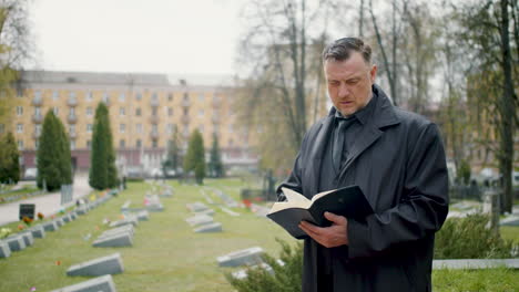 Man-In-Black-Raincoat-Looking-At-The-Horizon-In-A-Graveyard-And-Reading-A-Bible