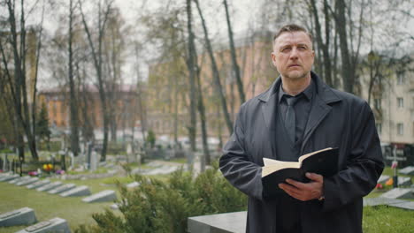 Man-In-Black-Raincoat-Looking-At-The-Horizon-In-A-Graveyard-And-Reading-A-Bible-1