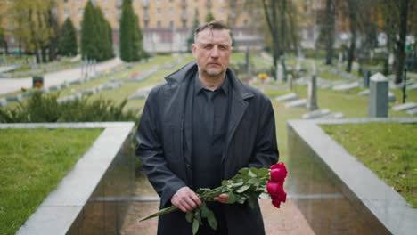 Front-View-Of-Man-In-Black-Raincoat-And-Suit-Holding-Roses-While-Walking-In-A-Graveyard