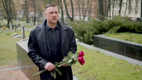 Man-In-Black-Raincoat-And-Suit-Holding-Roses-While-Walking-In-A-Graveyard