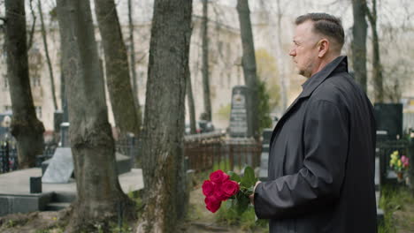 Side-View-Of-Man-In-Black-Raincoat-And-Suit-Holding-Roses-While-Walking-In-A-Graveyard