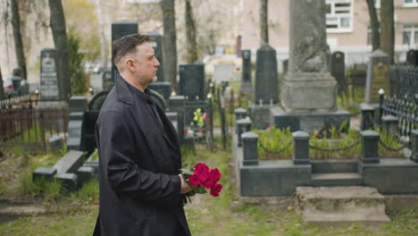 Side-View-Of-Man-In-Black-Raincoat-And-Suit-Holding-Roses-While-Walking-In-A-Graveyard-1
