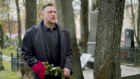 Man-In-Black-Raincoat-And-Suit-Holding-Roses-While-Walking-In-A-Graveyard-1