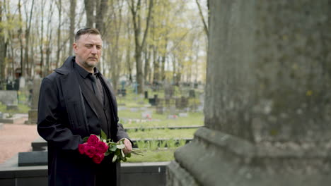 Man-In-Black-Raincoat-And-Suit-Holding-Red-Roses-Standing-In-Front-Of-A-Tombstone-In-A-Graveyard